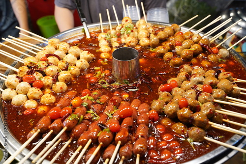 A large batch of Thai street food meatball skewers soaked and simmered in thick sweet, sour, and spicy red chili sauce on a big street market pan.