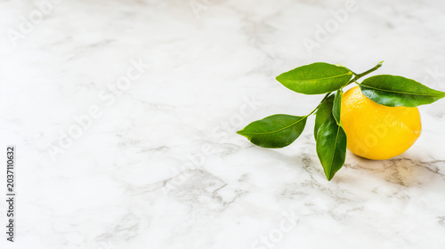 Fresh lemon with green leaves on a marble surface, showcasing vibrant yellow against a clean white background.