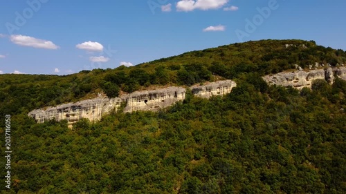 Aerial slow motion tracking shot of limestone cliff and forested hillside under clear blue sky
