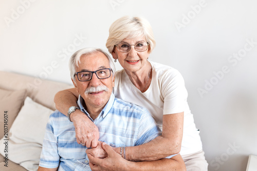 Happy senior couple portrait. Elderly people in love and healthy relationship, aging concept. Smiling mature man and elderly woman hugging and holding hands sitting on couch in bright living room.