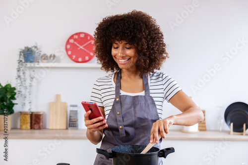 Canvastavla Happy young woman chef using mobile phone and cooking following online recipe in