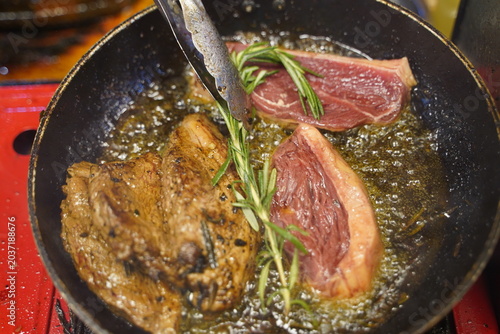 Close up of a beef steak being seared and fried in hot sizzling oil inside a pan, with fresh rosemary herb and metal tongs adjusting the food.