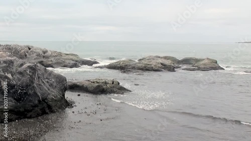 Gentle waves gracefully embrace the rugged coastline of Arctic Svalbard, Norway, on a calm, overcast day. Ancient rocks stand firm as the tide ebbs and flows, creating a serene, untamed landscape.