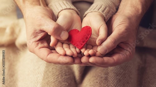 Adult and child hands holding a small red heart together over a warm knitted blanket