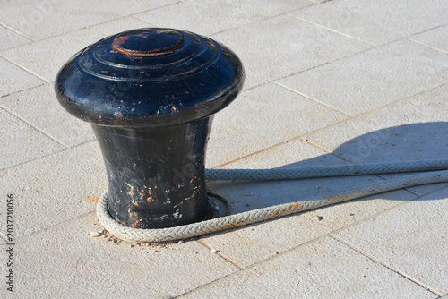 фотографія A heavy black metal mooring bollard with a secure rope wrapped around its base on a paved stone pier