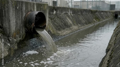 Sewage flowing from a rusted pipe into a polluted drainage channel with cloudy water reflecting the surrounding area