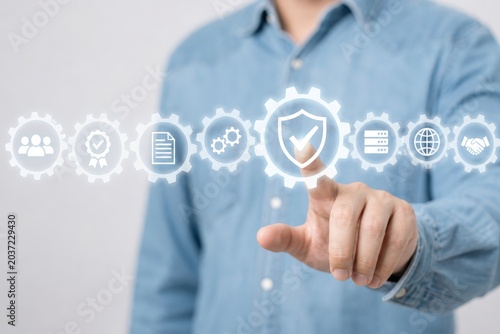 Man in a blue shirt presses a glowing icon of a shield with a checkmark within a series of interconnected gears representing security and quality assurance.