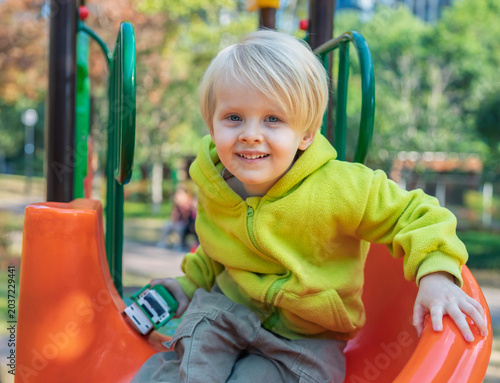 Cheerful toddler boy smiling while playing on a colorful playground slide in a bustling urban park in fall