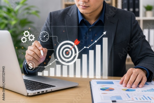 Man in a suit uses a stylus to point at a holographic chart showing upward trends, a target icon with an arrow, and gears.