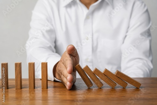 Man in a white shirt uses his hand to stop a row of falling dominoes, symbolizing risk management and crisis prevention in business.