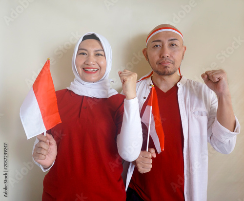Happy Asian Couple Clenched Fist Show Support For National Football Team While Holding Indonesian Little Flag. Concept Of Sports, Football Fans And Celebrating Independence Day