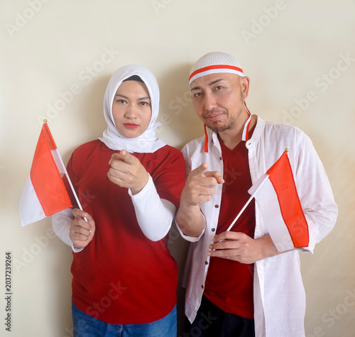 Serious Asian Couple Pointing At Camera Showing It's You Gesture While Holding Indonesian Flag. 