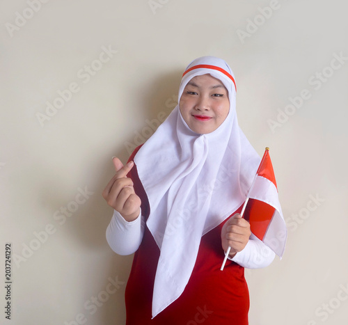 Asian Girl Wearing Headband And Hijab Showing Little Love Sign Using Finger While Holding Indonesian National Flag. Independence Day And Football Fans Concept