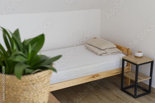 Bright attic bedroom with minimalist, wooden bed, white bed sheets, loft style bedside table and blurred, green plant in the foreground
