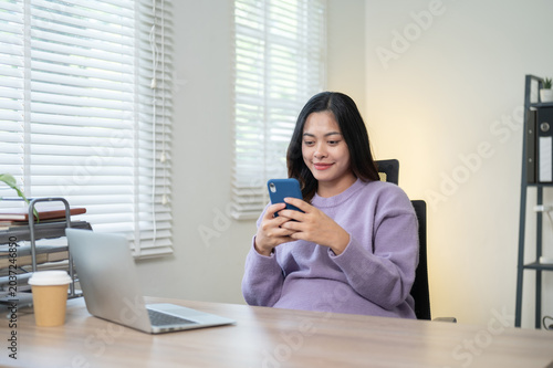 Pretty asian woman looking at phone beside laptop and coffee while sitting at wooden table in home.