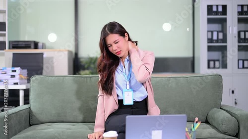 Tired Asian businesswoman suffering from office syndrome, rubbing her painful back, sore neck, and wrist while working.
