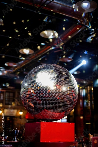A large mirrored disco ball on a red pedestal reflects colorful spotlights in a dark, atmospheric indoor event venue with exposed ceiling beams.