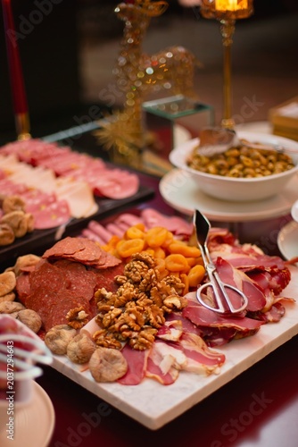 Charcuterie board loaded with sliced salami, cured meats, walnuts, dried figs, and apricots, served with tongs at an elegant buffet.