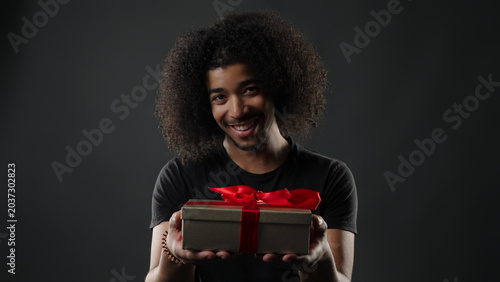 Smiling young man presenting a gift box with a red ribbon on a dark background