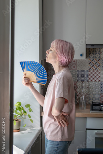 Weary middle aged woman standing in kitchen by window use handheld fan for relief from intense heat. Heatwave, no air conditioning, stifling indoor temperature. Female coping with high warmth at home.