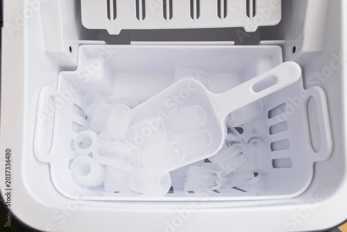 Overhead view inside modern white ice maker showing basket filled with clear bullet ice cubes and plastic scoop. 