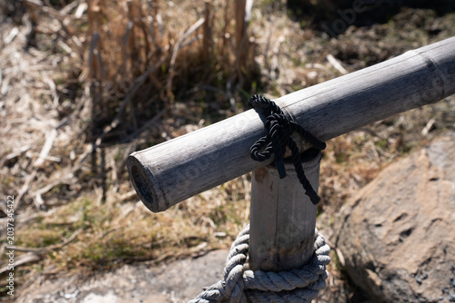 weathered bamboo pole secured to a wooden post with a coiled black rope and thick natural fiber line, resting on a sunlit rocky bank amid dry grasses rustic, textured beige-gray tones, negativespace