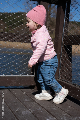 profile of a young toddler girl wearing a pink beanie and floral jacket, denim jeans and white sneakers, standing on a wooden dock by a lakeside, leaning against wire-mesh railing in bright sunlight