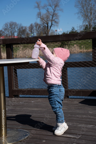 toddler in pink beanie and jacket standing on a wooden lakeside deck, reaching up to place a pastel bottle on a round table, sunny spring day with blue sky and bare trees, perspective, contrast