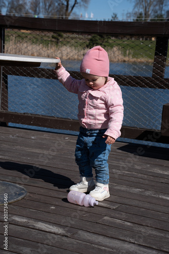 toddler in a pink beanie and patterned jacket stands on a wooden lakeside boardwalk beside a wire-fenced railing, gazing at a fallen pastel water bottle; sunny day, calm blue water and shadows