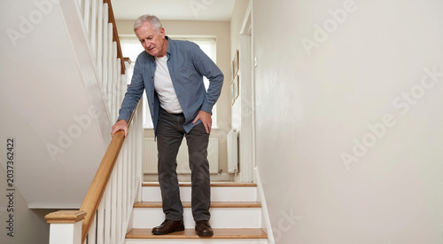 Senior man suffering from hip pain while descending stairs at home. Elderly male holding his painful joint due to osteoarthritis. Mobility restriction and skeletal degeneration concept