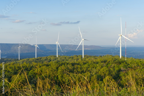 Panoramic view of wind turbines for renewable energy on a green forested hill with mountains under a bright evening sky.