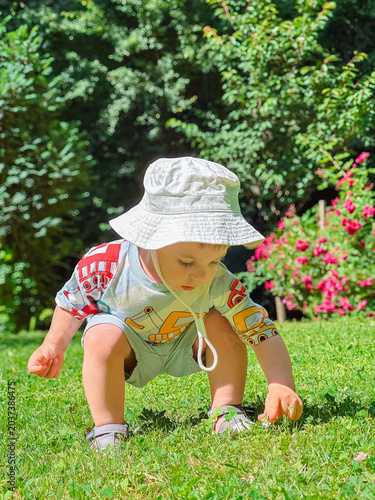 Toddler in sun hat squatting on green lawn in sunny garden