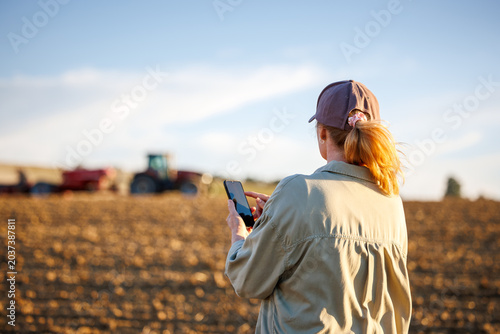 Woman farmer with smartphone monitoring smart farming cultivation while tractor and seeder prepare soil during agricultural work on field