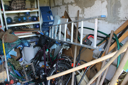 Cluttered garage interior filled with bicycles, ladders, tools, and various household items stacked haphazardly against a concrete wall