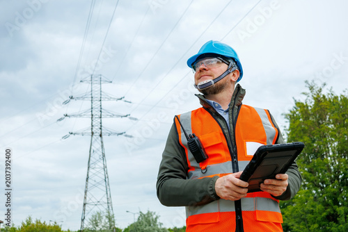 Portrait of a power engineer working near a high-voltage power line. Technician or inspector evaluates measures to prevent damage to the electrical grid infrastructure during a site inspection.
