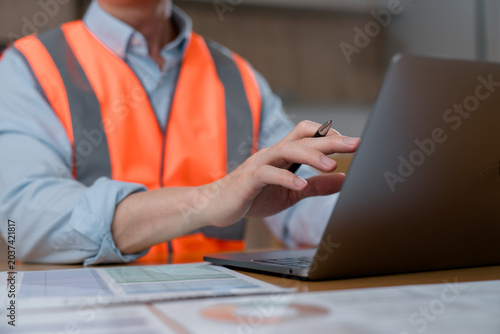 Canvastavla Close-up of an industrial worker wearing high visibility safety vest using laptop computer and reviewing documents in office or construction workplace environment