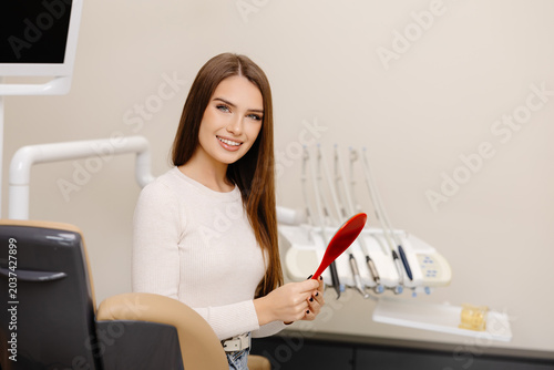 Pretty girl smiling while sitting in the dentist's chair, holding a mirror in her hands