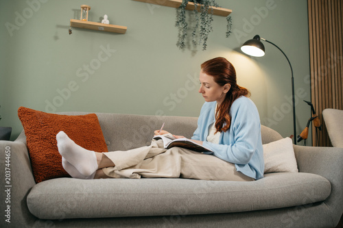 Obraz na płótnie Redhead woman relaxing on cozy sofa and writing notes in notebook at home during