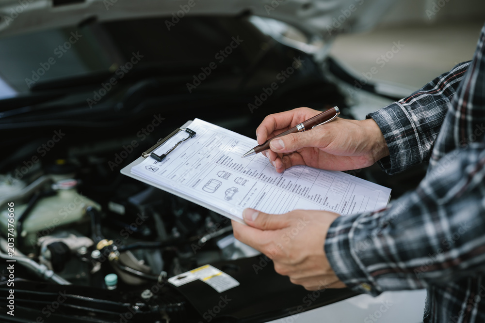Fototapeta premium Mechanic completing a vehicle inspection checklist under an open hood, documenting maintenance findings and evaluating engine condition for professional automotive service.