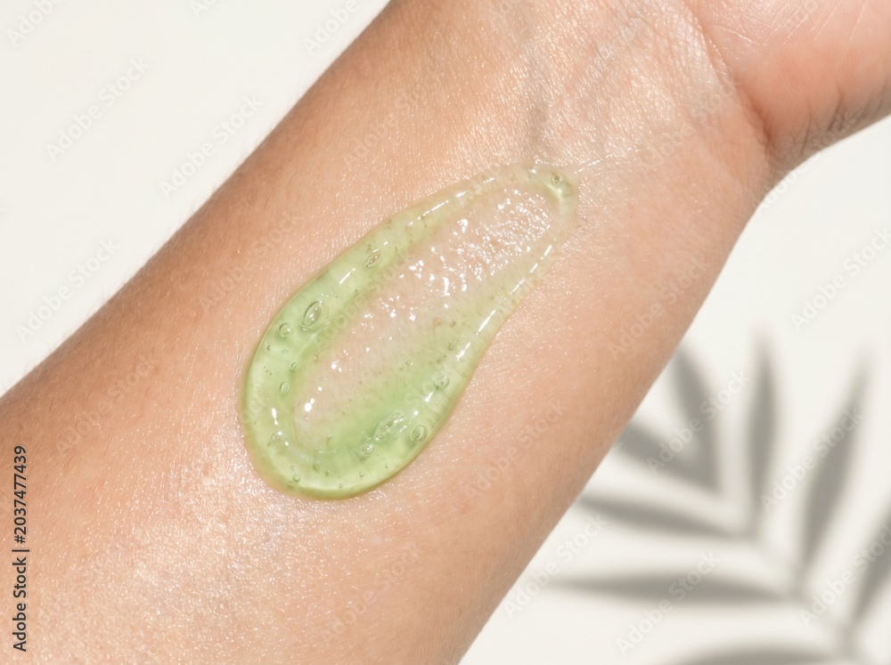 Fototapeta premium A dollop of clear green gel with small bubbles is swatched on a person's arm, with a blurred background of palm leaves.