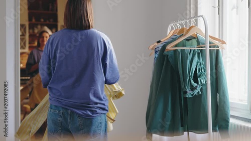 Adult woman choosing an outfit while looking in the mirror near a clothing rack at home. Morning routine, personal style, and everyday fashion decision in a bright modern apartment.