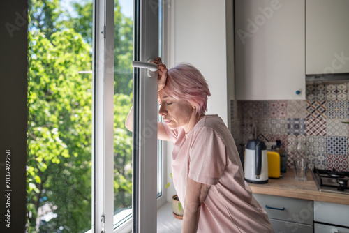 Bild auf Leinwand Tired woman standing near open window, leaning against frame while overheat, lack of air