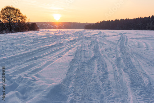 Deep snowmobile tracks crisscross a vast frozen field under a soft January sunset, creating textured patterns leading toward the warm golden horizon in a peaceful winter wilderness