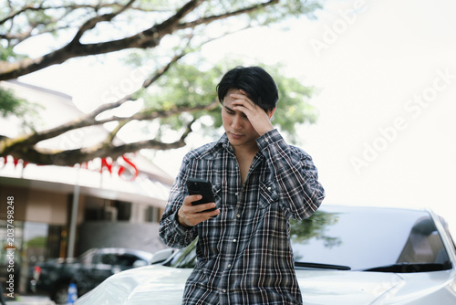 Obraz na plátne Worried young man looking at a smartphone beside a car, expressing financial stress, unexpected expenses, bad news, problem solving, and personal finance concerns
