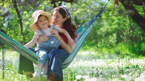 Happy young mother and her five-year-old daughter laughing and swinging in a hammock in a blooming summer garden. Warm family bonding, carefree childhood, outdoor lifestyle.