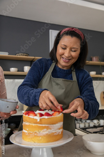 Family helping woman in gray apron decorating layered cake with strawberries on pedestal in kitchen