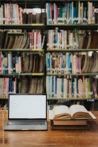 A simulated image of an empty laptop screen on a wooden table with a stack of books in a library, with a library as the backdrop.