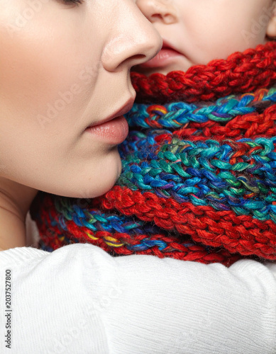 The mother wraps a warm knitted scarf around her child, showing only their hands and the colorful wool texture.
