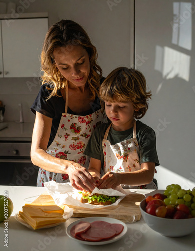 A mother helps her child prepare a school snack, hands visible wrapping a sandwich in wax paper.