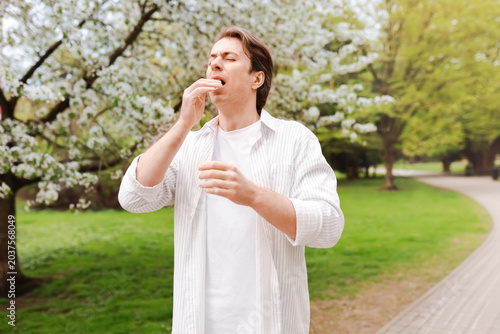 Fotografía A man suffering from seasonal allergy and hay fever sneezes in a nature park surrounded by blooming spring trees with pollen